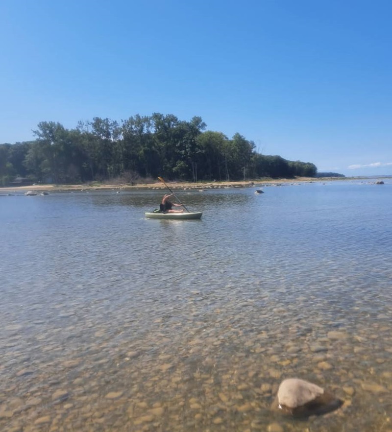 : Kayakers approaching Turnip Rock on Lake Huron, showing the limestone formation’s wide crown with trees and its eroded base surrounded by clear blue water.