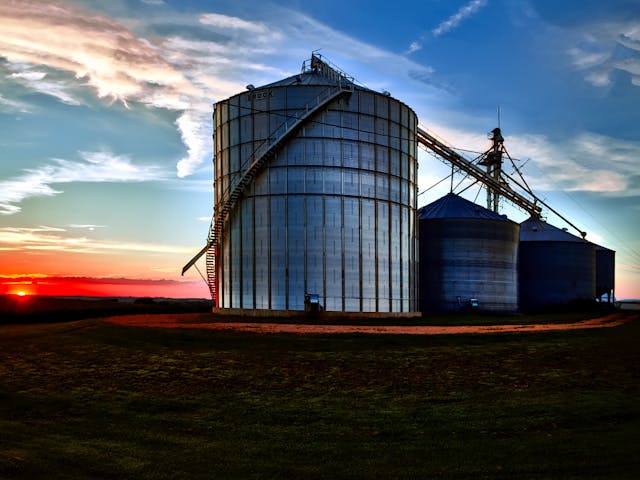 Large metal grain silos with conveyor system on farmland at sunset, silhouetted against a colorful evening sky.